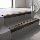 Square-edge aluminium stair nosing with a deep 56mm riser face in Satin Black, fitted along two vinyl-covered concrete steps in an office stairwell, with a Brown anti-slip PVC insert running the full width of each step