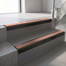 Square-edge aluminium stair nosing with a deep 56mm riser face in Satin Black, fitted along two vinyl-covered concrete steps in an office stairwell, with a Mid Brown anti-slip PVC insert running the full width of each step