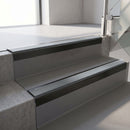 Square-edge aluminium stair nosing with a deep 56mm riser face in Satin Black, fitted along two vinyl-covered concrete steps in an office stairwell, with a Mid Grey anti-slip PVC insert running the full width of each step