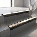 Square-edge aluminium stair nosing with a deep 56mm riser face in Satin Black, fitted along two vinyl-covered concrete steps in an office stairwell, with a Sahara anti-slip PVC insert running the full width of each step