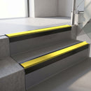 Square-edge aluminium stair nosing with a deep 56mm riser face in Satin Black, fitted along two vinyl-covered concrete steps in an office stairwell, with a Yellow anti-slip PVC insert running the full width of each step