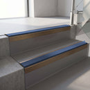 Square-edge aluminium stair nosing with a deep 56mm riser face in Satin Bronze, fitted along two vinyl-covered concrete steps in an office stairwell, with a Blue anti-slip PVC insert running the full width of each step
