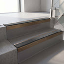 Square-edge aluminium stair nosing with a deep 56mm riser face in Satin Bronze, fitted along two vinyl-covered concrete steps in an office stairwell, with a Dark Grey anti-slip PVC insert running the full width of each step