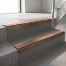 Square-edge aluminium stair nosing with a deep 56mm riser face in Satin Bronze, fitted along two vinyl-covered concrete steps in an office stairwell, with a Mid Brown anti-slip PVC insert running the full width of each step