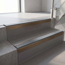 Square-edge aluminium stair nosing with a deep 56mm riser face in Satin Bronze, fitted along two vinyl-covered concrete steps in an office stairwell, with a Mid Grey anti-slip PVC insert running the full width of each step