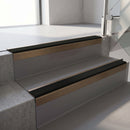 Square-edge aluminium stair nosing with a deep 56mm riser face in Satin Bronze, fitted along two vinyl-covered concrete steps in an office stairwell, with a Premium Black anti-slip PVC insert running the full width of each step