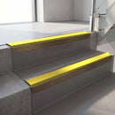 Square-edge aluminium stair nosing with a deep 56mm riser face in Satin Bronze, fitted along two vinyl-covered concrete steps in an office stairwell, with a Yellow anti-slip PVC insert running the full width of each step