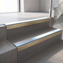 Square-edge aluminium stair nosing with a deep 56mm riser face in Satin Gold, fitted along two vinyl-covered concrete steps in an office stairwell, with an Aura anti-slip PVC insert running the full width of each step