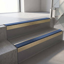 Square-edge aluminium stair nosing with a deep 56mm riser face in Satin Gold, fitted along two vinyl-covered concrete steps in an office stairwell, with a Blue anti-slip PVC insert running the full width of each step