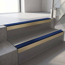 Square-edge aluminium stair nosing with a deep 56mm riser face in Satin Gold, fitted along two vinyl-covered concrete steps in an office stairwell, with a Dark Blue anti-slip PVC insert running the full width of each step