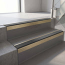Square-edge aluminium stair nosing with a deep 56mm riser face in Satin Gold, fitted along two vinyl-covered concrete steps in an office stairwell, with a Dark Grey anti-slip PVC insert running the full width of each step