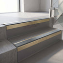 Square-edge aluminium stair nosing with a deep 56mm riser face in Satin Gold, fitted along two vinyl-covered concrete steps in an office stairwell, with a Mid Grey anti-slip PVC insert running the full width of each step