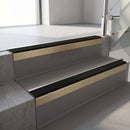 Square-edge aluminium stair nosing with a deep 56mm riser face in Satin Gold, fitted along two vinyl-covered concrete steps in an office stairwell, with a Premium Black anti-slip PVC insert running the full width of each step