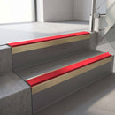 Square-edge aluminium stair nosing with a deep 56mm riser face in Satin Gold, fitted along two vinyl-covered concrete steps in an office stairwell, with a Red anti-slip PVC insert running the full width of each step