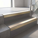Square-edge aluminium stair nosing with a deep 56mm riser face in Satin Gold, fitted along two vinyl-covered concrete steps in an office stairwell, with a Sahara anti-slip PVC insert running the full width of each step