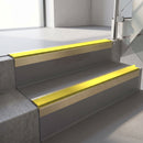 Square-edge aluminium stair nosing with a deep 56mm riser face in Satin Gold, fitted along two vinyl-covered concrete steps in an office stairwell, with a Yellow anti-slip PVC insert running the full width of each step