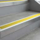 Bright Polished deep-riser square-edge aluminium stair nosing fitted along two bare concrete steps, 62mm tread plate and 56mm front face, with a Yellow anti-slip insert running the full width of each step