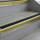 Mirrorflect Gold deep-riser square-edge aluminium stair nosing fitted along two bare concrete steps, 62mm tread plate and 56mm front face, with a Dark Blue anti-slip insert running the full width of each step