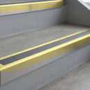 Satin Gold deep-riser square-edge aluminium stair nosing fitted along two bare concrete steps, 62mm tread plate and 56mm front face, with a Mid Grey anti-slip insert running the full width of each step