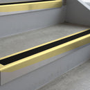 Satin Gold deep-riser square-edge aluminium stair nosing fitted along two bare concrete steps, 62mm tread plate and 56mm front face, with a Premium Black anti-slip insert running the full width of each step