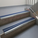 Bright Polished aluminium stair nosing fitted along two concrete steps in an office stairwell, with twin Blue anti-slip inserts running the full width of each step