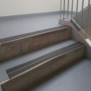 Dark Bronze aluminium stair nosing fitted along two concrete steps in an office stairwell, with twin Dark Grey anti-slip inserts running the full width of each step