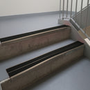 Dark Bronze aluminium stair nosing fitted along two concrete steps in an office stairwell, with twin Premium Black anti-slip inserts running the full width of each step