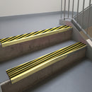 Satin Gold aluminium stair nosing fitted along two concrete steps in an office stairwell, with twin Hazard black and yellow anti-slip inserts running the full width of each step