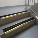 Satin Gold aluminium stair nosing fitted along two concrete steps in an office stairwell, with twin Premium Black anti-slip inserts running the full width of each step