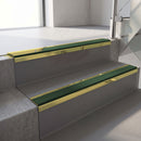 Mirrorflect Gold mirror-finish aluminium stair nosing fitted along two concrete steps in an office stairwell, with a Dark Green anti-slip insert running the full width of each step