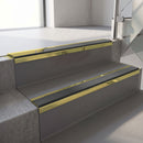 Mirrorflect Gold mirror-finish aluminium stair nosing fitted along two concrete steps in an office stairwell, with a Dark Grey anti-slip insert running the full width of each step