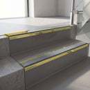 Mirrorflect Gold mirror-finish aluminium stair nosing fitted along two concrete steps in an office stairwell, with a Mid Grey anti-slip insert running the full width of each step