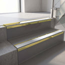 Mirrorflect Gold mirror-finish aluminium stair nosing fitted along two concrete steps in an office stairwell, with a Polar Grey anti-slip insert running the full width of each step
