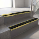 Mirrorflect Gold mirror-finish aluminium stair nosing fitted along two concrete steps in an office stairwell, with a Premium Black anti-slip insert running the full width of each step
