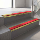 Mirrorflect Gold mirror-finish aluminium stair nosing fitted along two concrete steps in an office stairwell, with a Red anti-slip insert running the full width of each step
