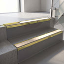 Mirrorflect Gold mirror-finish aluminium stair nosing fitted along two concrete steps in an office stairwell, with a Sahara anti-slip insert running the full width of each step