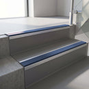 Square-edge aluminium stair nosing fitted along two concrete steps in an office stairwell, with a Blue anti-slip insert running the full width of each step