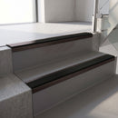 Dark Bronze aluminium stair nosing fitted along two concrete steps in an office stairwell, with a Premium Black anti-slip insert running the full width of each step