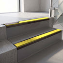 Dark Bronze aluminium stair nosing fitted along two concrete steps in an office stairwell, with a Yellow anti-slip insert running the full width of each step