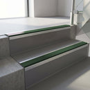 Square-edge aluminium stair nosing fitted along two concrete steps in an office stairwell, with a Dark Green anti-slip insert running the full width of each step