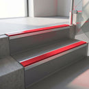 Square-edge aluminium stair nosing fitted along two concrete steps in an office stairwell, with a Red anti-slip insert running the full width of each step