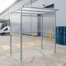 Galvanised smoking shelter installed on a concrete warehouse yard beside two yellow safety bollards, a stack of blue plastic pallets and a white van at the loading bay