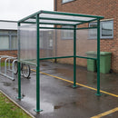Moss Green outdoor smoking shelter bolted to wet tarmac in a red-brick school courtyard, with a bicycle on a galvanised cycle stand to the left and two green wheelie bins against the wall