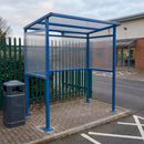 Traffic Blue smoking shelter on block paving at the edge of a staff car park, with a grey pedestal litter bin to the left, green palisade fencing and hedge behind and white bay markings in front