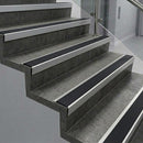Raked back nosings with smooth black inserts on dark concrete steps beside a glass balustrade
