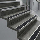 Raked back nosings with smooth black inserts on dark concrete steps beside a glass balustrade