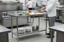 Chef preparing fresh vegetables on a 1200 x 600mm stainless steel prep table in a busy commercial kitchen, with mixing bowl, plates and storage trays on the galvanised undershelf