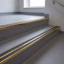 Square edge aluminium stair nosing with twin dark charcoal Mor Grip inserts in mirrorflect gold finish fitted on a school corridor stair finished in grey sheet vinyl