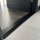 Black anodised aluminium door threshold ramp fitted across a doorway where dark charcoal LVT meets pale porcelain tile in a kitchen