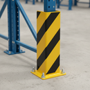 Close-up of a yellow powder-coated steel racking upright protector's black chevron stripes and floor-bolted base plate on a warehouse pallet racking upright